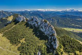 Luftaufnahme eines bewaldeten Gebirgsgrats mit schroffen Felsen und dahinter sichtbaren, schneebedeckten Alpenbergen unter klarem, blauem Himmel.