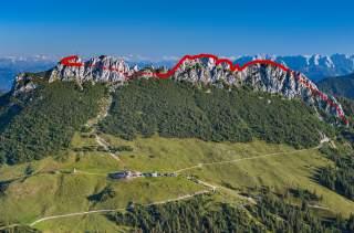 Bergpanorama mit grün bewachsenen Hängen und schroffen Felsgipfeln, auf denen eine rote Linie mit Punkten einen Wanderweg markiert. Im Vordergrund sind Wiesen, Gebäude und Wege zu sehen.