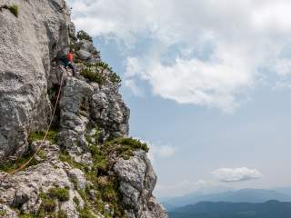 Eine Person klettert mit Helm und Seil an einer felsigen Bergwand, im Hintergrund sind bewaldete Berge und ein bewölkter Himmel zu sehen.
