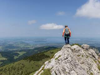 Wanderer mit Helm und Rucksack steht auf einem felsigen Gipfel und blickt auf ein weites Tal mit Seen und Dörfern bei klarem Himmel.