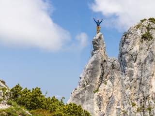 Ein Kletterer steht auf einem spitzen Felsgipfel mit erhobenen Armen, gesichert durch ein Seil, umgeben von Felsen und blauem Himmel.