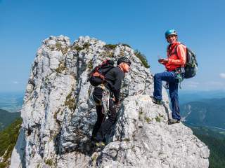 Zwei Kletterer mit Helmen und Rucksäcken sichern sich an einem felsigen Gipfel unter klarem Himmel, einer hält eine Karte.