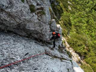 Kletterer mit Helm und Rucksack steigt an einer steilen Felswand, gesichert durch ein rotes Seil, mit grün bewachsenem Gelände im Hintergrund.