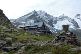 Berghütte in alpiner Landschaft vor schneebedeckten Bergen, im Vordergrund ein Steinmandl