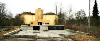 Verlassenes, gelbes Industriegebäude mit runden Silos, umgeben von überwachsenem Gelände und kahlen Bäumen.