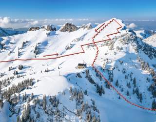 Luftaufnahme eines schneebedeckten Berges mit eingezeichneter roter Route, die im Zickzack auf den Gipfel führt.