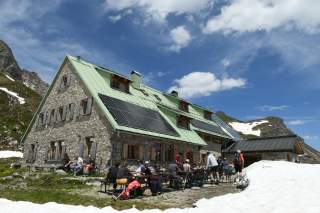 Auf Hüttenterrasse genießen Menschen die Sonne, im Vordergrund liegt noch Schnee