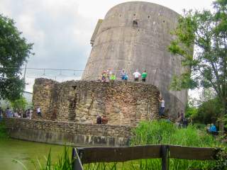 An einem schiefen Hochbunker klettern einige Menschen, Kinder wie Erwachsene. Davor eine alte Natursteinmauer.