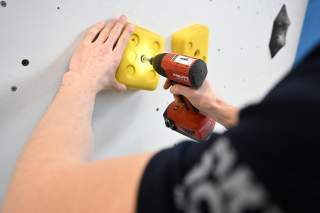 Die linke Hand hält einen Klettergriff, mit der rechten Hand wird er mit einem Akkuschrauber angeschraub.