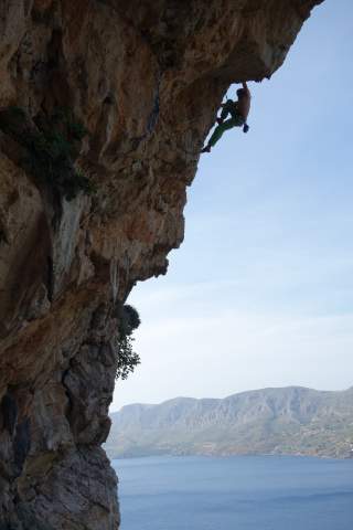Ein Kletterer hängt an einer steilen Felswand über dem Meer, im Hintergrund sind Berge und Wasser zu sehen.