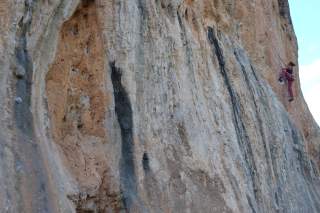 Person klettert an einer steilen, rötlich-grauen Felswand mit Sicherungsseil.