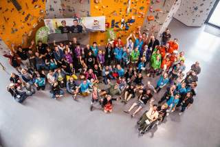 Große Gruppe lachender Menschen, darunter auch eine Person im Rollstuhl, versammelt in einer Kletterhalle vor einer Boulderwand, viele heben die Arme und blicken zur Kamera. Im Hintergrund hängt ein Banner der Special Olympics Landesspiele.