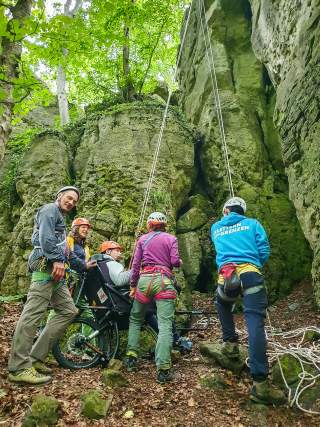 Fünf Personen in Kletterausrüstung heben gemeinsam einen Mann in einem Spezialrollstuhl in einer felsigen Waldlandschaft, um ihn beim Klettern zu unterstützen. Seile sind an den Felsen befestigt.