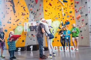 Menschen feiern eine Siegerehrung vor einer Kletterwand, eine Person wird umarmt, andere halten Urkunden und Medaillen in der Hand.