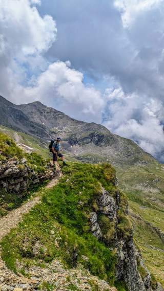 Ein Wanderer steht auf einem schmalen Wanderweg in den Alpen. Der Himmel voller Wolken.