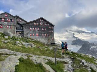 Große Berghütte mit rot-weißen Fensterläden steht auf einer Almwiese, zwei Wanderer mit Rucksäcken gehen auf einem Pfad, im Hintergrund schneebedeckte Berge und Gletscher.