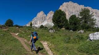 Bergsteiger wandert über Wiesenlandschaft am Fuße eines Felsmassivs.