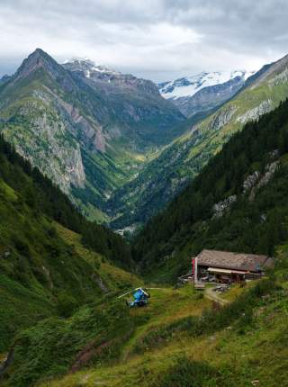 Blick auf ein grünes Bergtal mit bewaldeten Hängen, einer kleinen Berghütte und einem blauen Hubschrauber auf einer Wiese, im Hintergrund schneebedeckte Berggipfel unter bewölktem Himmel.