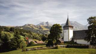 Ein Bergdorf. Rechts eine alte Kirche, im Bild eine Schweizer Fahne, im Hintergrund hohe Berge.