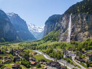 Panorama eines malerischen Tals mit einem Fluss, Wasserfall, traditionellen Häusern und schneebedeckten Bergen im Hintergrund.