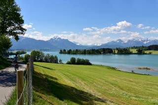 Alpenlandschaft mit einem See im Vordergrund und Wiesen, auf dem Weg links ein Radler.