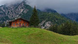 Kleine Holzalm vor einem felsigen Berg mit Wald, der von Nebel verdeckt ist.