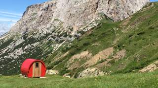 Grüne Wiese, darauf steht eine kleine rote Notunterkunft (Biwak) wie ein kleines Holzfass. Im Hintergrund Felsflanken oberhalb der Baumgrenze.