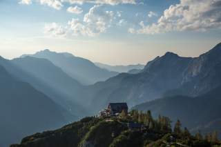 Berglandschaft mit nebligen Tälern und Sonnenstrahlen, die auf eine Berghütte auf einem bewaldeten Gipfel fallen, unter einem blauen Himmel mit vereinzelten Wolken.