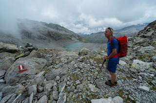 Wanderer mit rotem Rucksack steht auf einem steinigen Gebirgspfad, im Hintergrund ein türkisfarbener Bergsee und wolkenverhangene Berge.