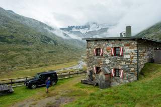 Steinhaus mit rot-weißen Fensterläden in den Bergen, davor ein schwarzer Geländewagen und eine Person mit Wanderausrüstung; im Hintergrund grüne Hänge, Nebel und schneebedeckte Gipfel.