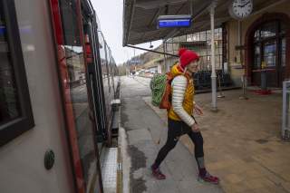 Frau mit rotem Wollmütze und gelber Jacke steigt mit Kletterrucksack aus einem Zug am Bahnhof Tegernsee aus.