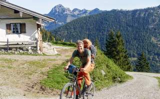 Ein Mann mit Rucksack fährt mit dem Fahrrad auf einem Schotterweg in den Bergen, links steht eine Berghütte, im Hintergrund bewaldete Hügel und Gipfel.