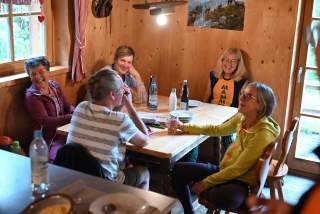 Fünf Personen sitzen lachend an einem Holztisch in einem gemütlichen, mit Holz verkleideten Raum. Auf dem Tisch stehen Getränkeflaschen, Gläser und Teller, während draußen durch das Fenster Tageslicht hereinfällt.