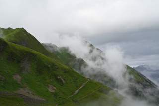 Grüne, nebelverhangene Berglandschaft unter bewölktem Himmel mit sichtbaren Wanderwegen.