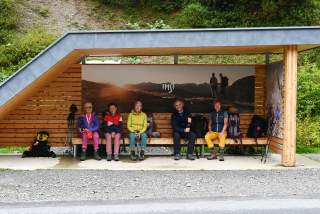 Fünf Personen in bunter Outdoorbekleidung sitzen auf einer Holzbank in einer überdachten Bushaltestelle, umgeben von Wanderstöcken und Rucksäcken.