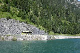 Ein gelber Bus fährt auf einer Straße an einem See entlang, umgeben von steilen Felswänden und dichtem Nadelwald.