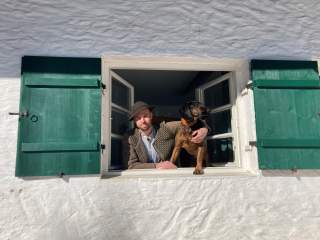 Mann und Hund lehnen sich aus einem Fenster mit grünen Holzläden.