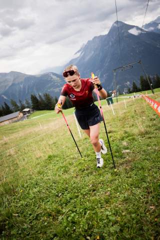 Eine Athletin käuft mit Trailrunstöcken eine grüne Wiese hoch. Sie trägt ein rotes T-Shirt und eine schwarze Hose. Im Hintergrund sieht man Berge, viele Wolken und eine Liftanlage.