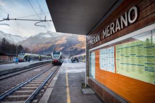Züge am Bahnhof Meran, im Vordergrund eine Fahrplantafel, im Hintergrund die Alpen.
