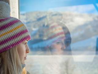 Frau mit bunter Mütze blickt nachdenklich aus einem Fenster, ihre Spiegelung ist auf dem Glas sichtbar. Draußen sind verschwommene Berglandschaften zu erkennen.