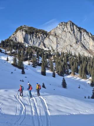 Drei Personen in Skitourenausrüstung stehen im Skitourengelände, vor ihnen bauen sich zwei Felsgipfel auf.