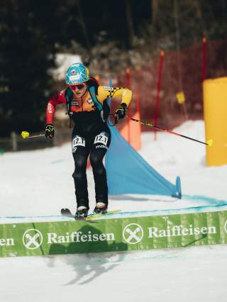 Eine Athletin sprint über eine kleine Chance in der Abfahrt. Sie ist gerade in der Luft. Blick ist fokussiert. Im Hintergrund sieht man ein blaues Abfahrtstor und Absperrungszäune.