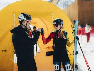 ein Athlet im Mantel und eine Athletin im Rennanzug lachen und klatschen sich ab vor dem Start. Hinter ihnen ein gelbes Zelt.