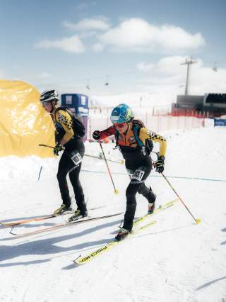 zwei Athleten, die gerade in der Wechselzone sind. Im Vordergrund ist eine Athletin, die dynamisch nach vorne läuft, während der Athlet im Hintergrund bremst und zur Zeit geht.