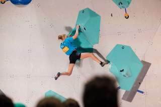 Frau klettert an einer künstlichen Boudlerwand vor Publikum