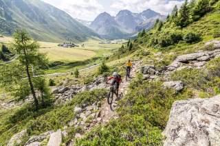 Zwei Mountainbiker fahren auf einem Wanderweg durch ein Bergtal.