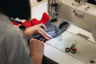 Person näht mit Nähmaschine dunkelblauen Stoff mit rotem Rand, neben der Maschine liegt eine kleine Schere.