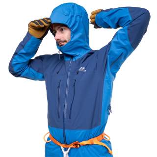 Mann zieht blaue Outdoorjacke mit Kapuze und Klettergurt an, trägt braune Handschuhe.
