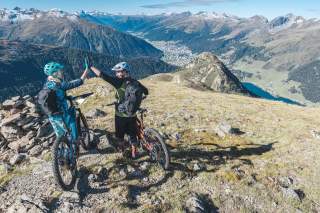 Zwei Mountainbiker auf einem Berggipfel mit Blick auf Davos schlagen ein.