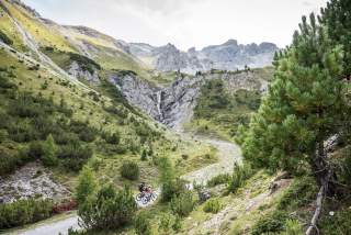 Zwei Mountainbiker fahren durch ein Bergtal in Richtung eines Wasserfalls.